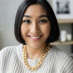 Portrait of a cheerful young woman wearing a gold necklace and sweater indoors with a blurred background.